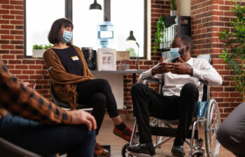 Man with disability talking at group therapy session with people having alcohol addiction. Patients wearing face masks and attending aa meeting to have discussion with psychologist.
