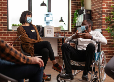 Man with disability talking at group therapy session with people having alcohol addiction. Patients wearing face masks and attending aa meeting to have discussion with psychologist.